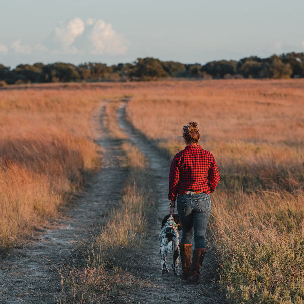 Person walking a dog on a dirt path through a field with trees in the background